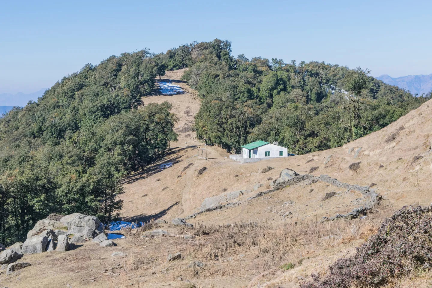 A small white building with a green roof on a dry, brown ridge, with dark green woods on both sides and a few patches of snow on the ground; the photographer calls it the clearing just before the temple.