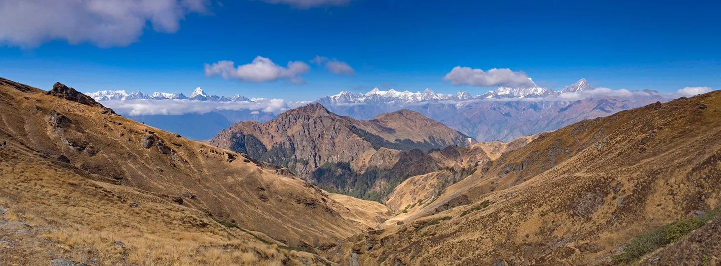A wide autumn view from Kuari Pass: golden grass ridges and a rocky spur falling into a V-shaped valley, a band of cloud, and a long serrated line of snow peaks on the horizon under a deep blue sky.