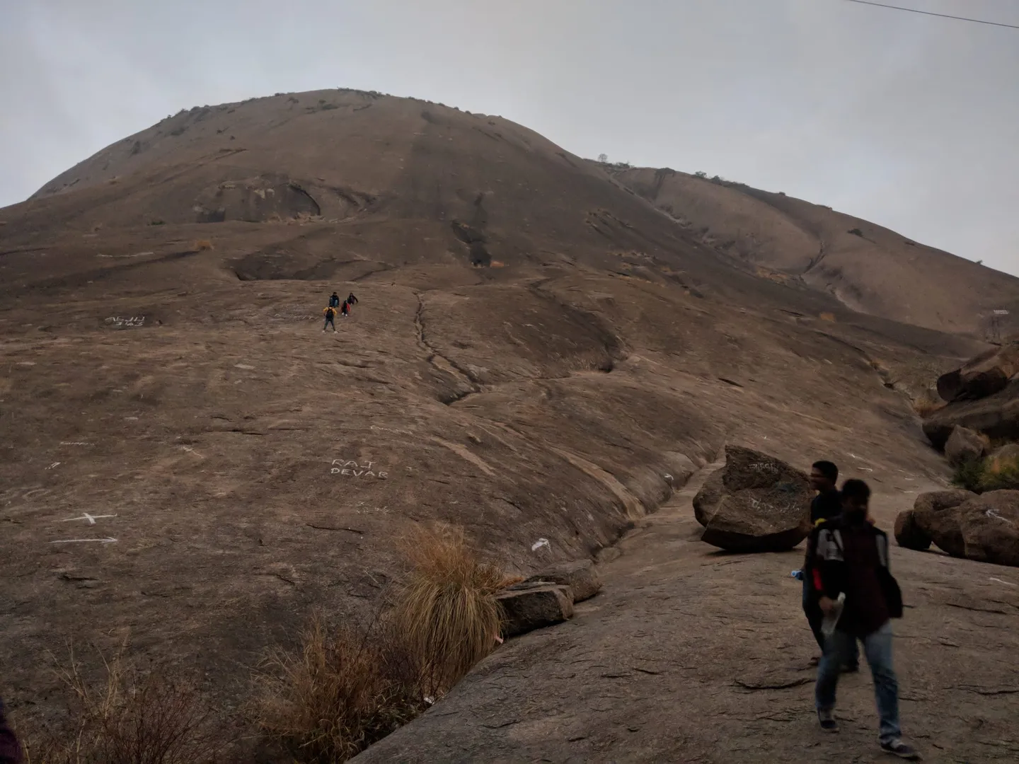 The climbing surface itself, under flat grey early-morning cloud: an unbroken sheet of granite rising at a steady angle towards the dome, with a faint darker line worn up the middle for a path and white painted names and arrows daubed on the rock. Two walkers in jeans and trainers are coming down in the foreground and three more are small dots high on the slope. No steps, no railing and no shade anywhere in the frame.