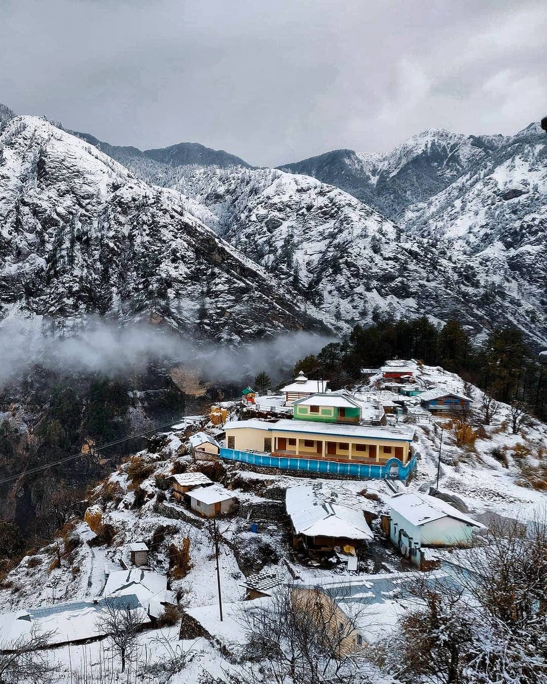 Sankri village under fresh snow, seen from above — pastel-roofed houses stacked on a hillside with low cloud in the forested valley behind. The roadhead where the expedition assembles.