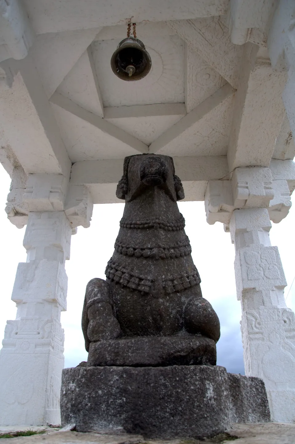 The Nandi at the top, photographed from directly behind and below, so its back, hump and haunches fill the frame and the head is seen from the rear with the face not visible: a dark, coarse, lichen-flecked stone bull on a plain rough plinth, three rows of carved bell-and-bead garlands running across its back and shoulders. It sits inside a small pavilion whose pillars and ceiling are thickly whitewashed over shallow carved rosettes, with a dark brass bell on a rusted chain above it and plain pale sky between the pillars.