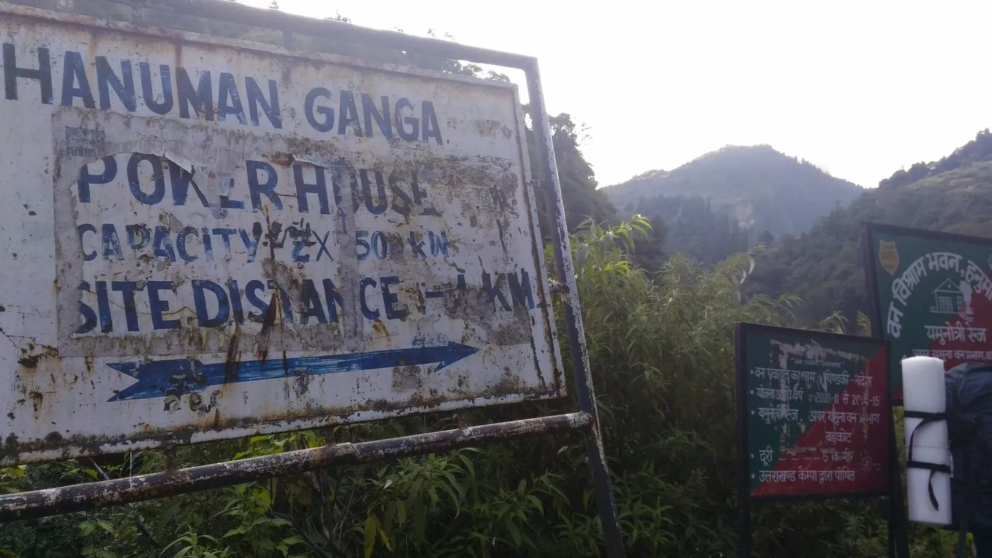 Beside the Hanuman Ganga near the trailhead on a September morning: a weathered white power-house sign with an arrow, and a green forest-department board in Hindi, in front of forested hills.