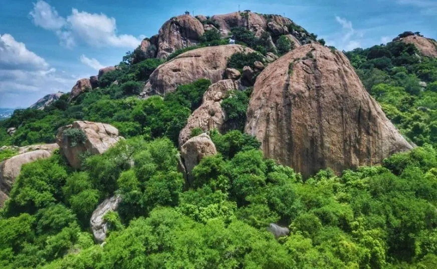 Kunagalu Betta from the air, rounded domes of bare brown granite rising out of dense green scrub and low trees, a small structure standing on the skyline at the summit and the plain hazy beyond the left shoulder of the hill.