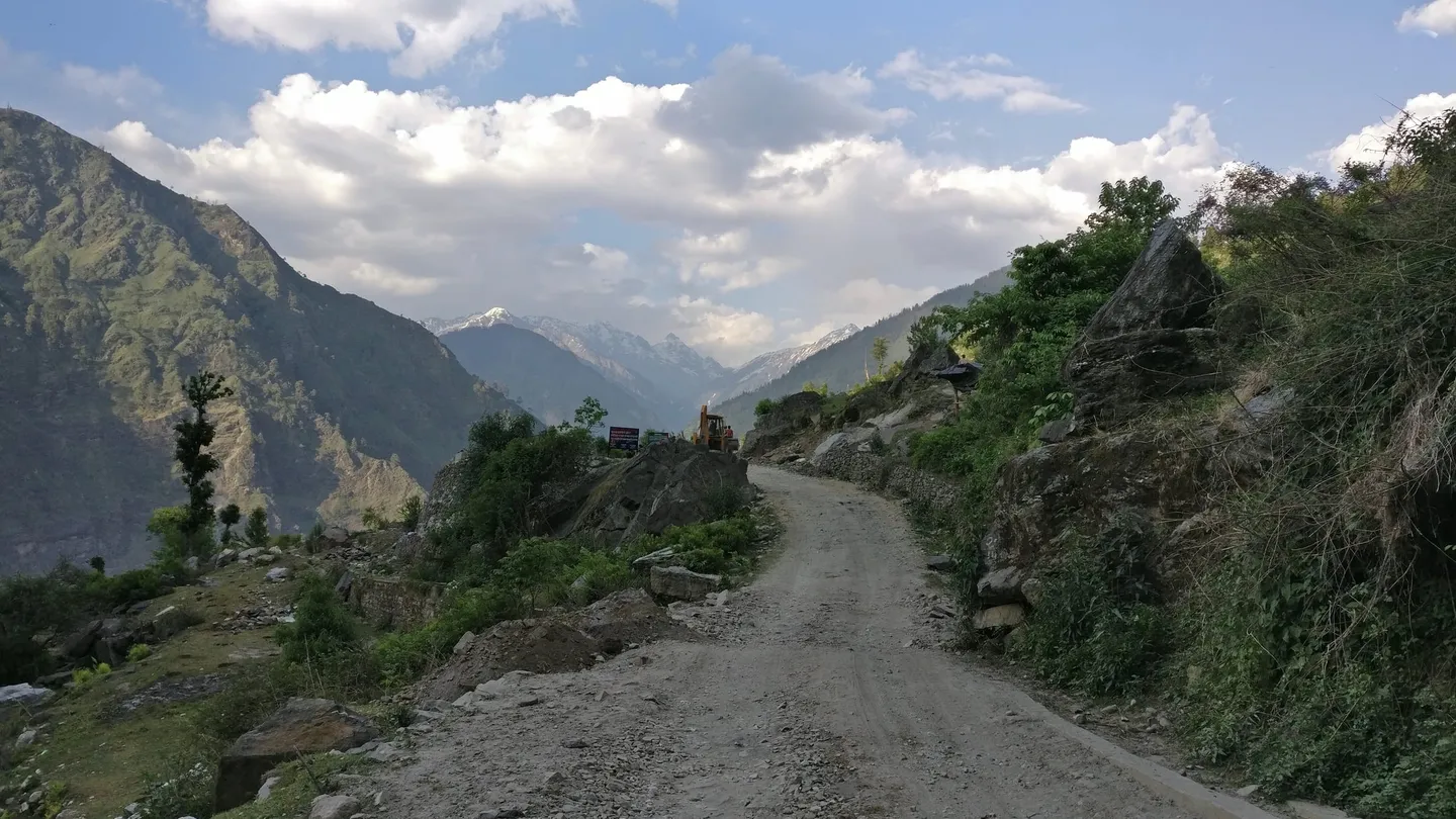 The unsurfaced road to Sankri, cut into a forested hillside with an excavator working on it and a distant snow range ahead — the last hours of the long drive from Dehradun to the trailhead.