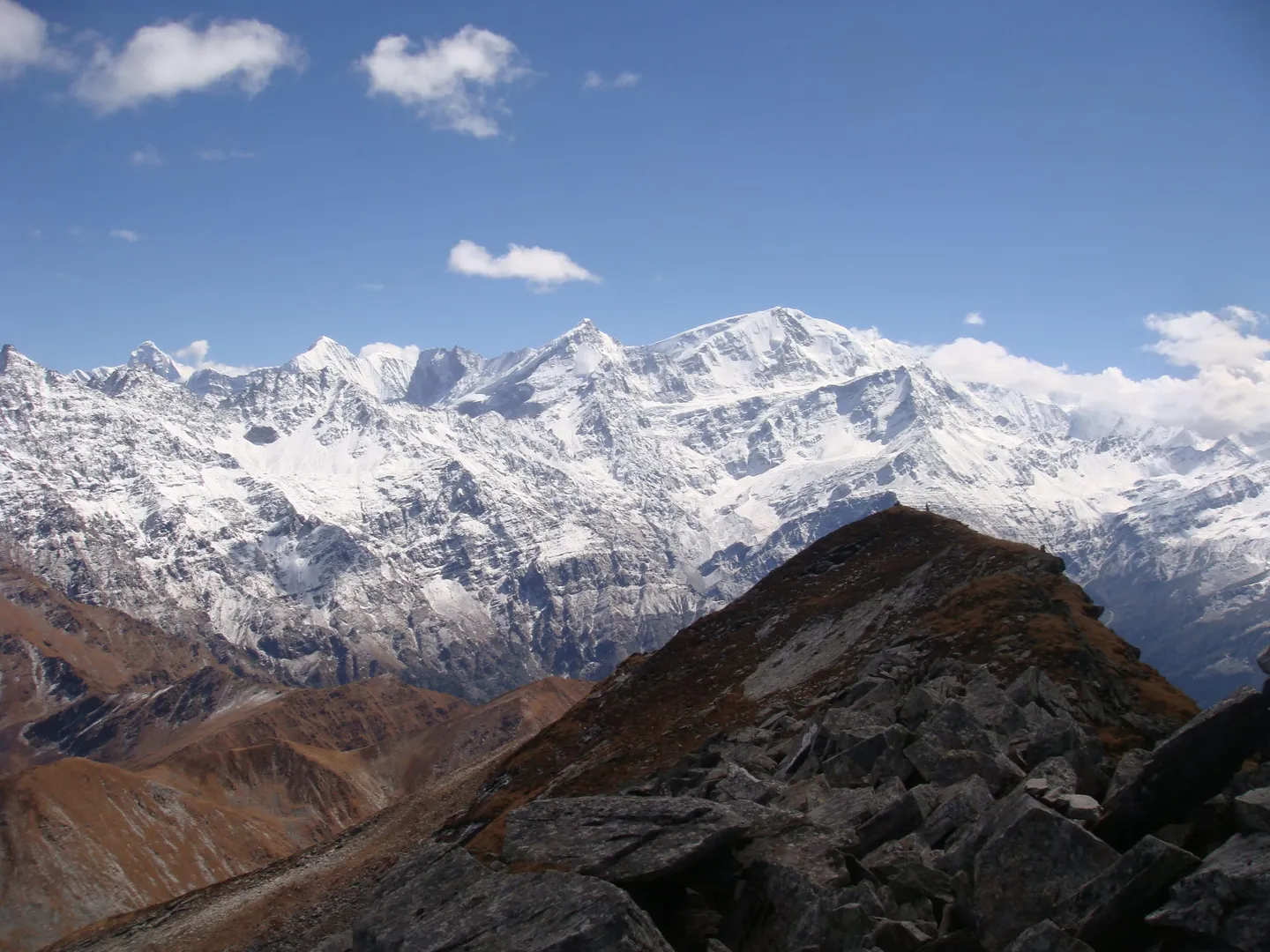A view from the Pangarchulla summit area: a bare brown, boulder-strewn ridge with a few small figures on its crest, and a broad snow-covered massif behind, under a deep blue sky with scattered cloud.