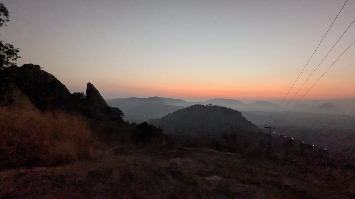 Dawn over the Ramanagara plain from Kunagalu Betta, a tall fin of granite standing in silhouette on the left and low hills receding into haze under an orange horizon.