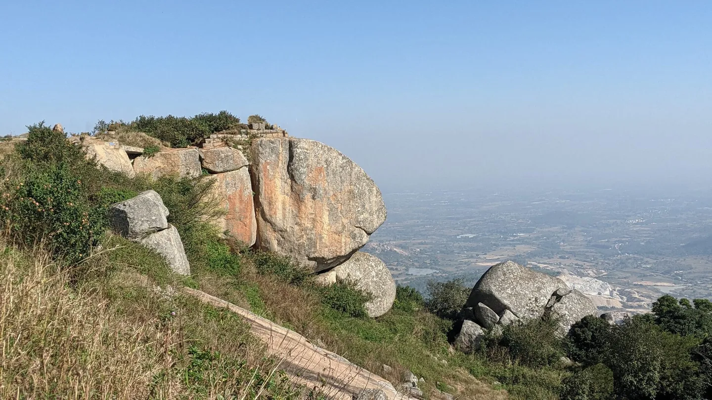 Near the top of Skandagiri, looking out over the edge: a cracked orange-grey granite outcrop with a detached block leaning against it, dry grass and thorn scrub in the foreground, and the hazy plain far below with fields, scattered villages, a pale quarry scar and a small reservoir. Photographed on the summit in April, in late-morning haze, not on the sunrise walk.