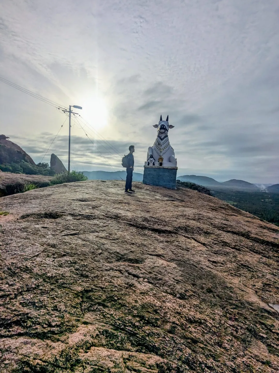 A white-painted Nandi bull on a stone plinth on the bare granite summit of Kunagalu Betta, a walker standing beside it and the hills of the Ramanagara plain behind.