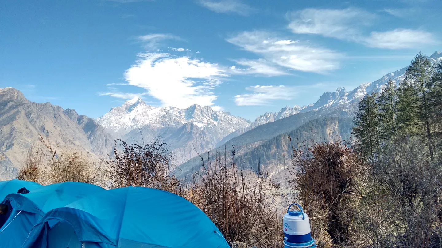 A December camp view that the photographer's description places on a trek to Kuari Pass: blue dome tents and a white water jug in the foreground, dry brown shrubs and a few pines, and a long line of snow-covered mountains under a blue sky with thin cloud.