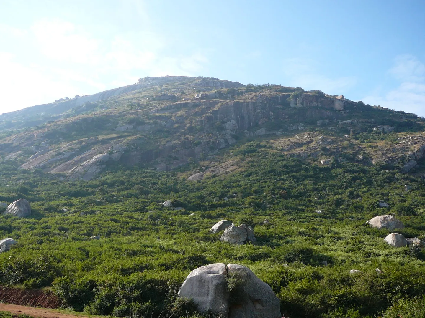The hill in daylight from close to its base: a red-earth track at the bottom left, then a wide apron of green scrub studded with rounded granite boulders, rising into bare rock slabs and a rocky summit ridge with a small structure just breaking the skyline. Low morning sun from the left, not the pre-dawn dark. The photograph is from December 2007 and carries no location data, so it shows the lower slopes of the hill rather than an identified point on the route.
