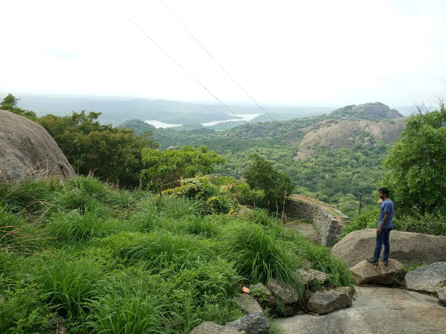 A hillside in monsoon green — tussock grass and scrub over granite boulders, a low dry-stone wall running across the slope on the right, a rounded granite dome opposite and a sheet of reservoir water far below. One person stands on a slab of rock for scale, and a power line crosses the white sky. Taken on Savanadurga in June, about 900 m from the point the Forest Department's own map pins for this trek — but up the neighbouring monolith, not on the Bidarakatte path.