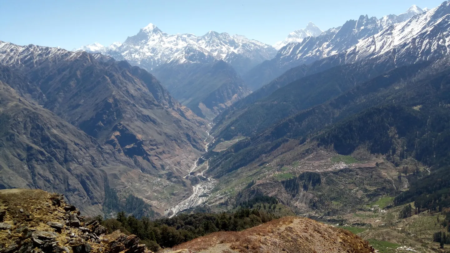 A spring view near Kuari Pass, taken in April: a deep valley with a river bed and terraced fields and villages on the far slope, dry grass ridges in the foreground, and snow mountains on the skyline under a clear sky.