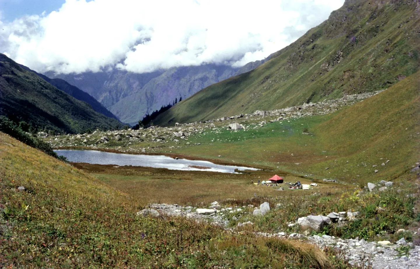 Ruinsara Tal on the approach — a shallow lake in a wide grassy valley strewn with boulders, a single orange tent pitched on the shore, and cloud filling the head of the valley beyond.
