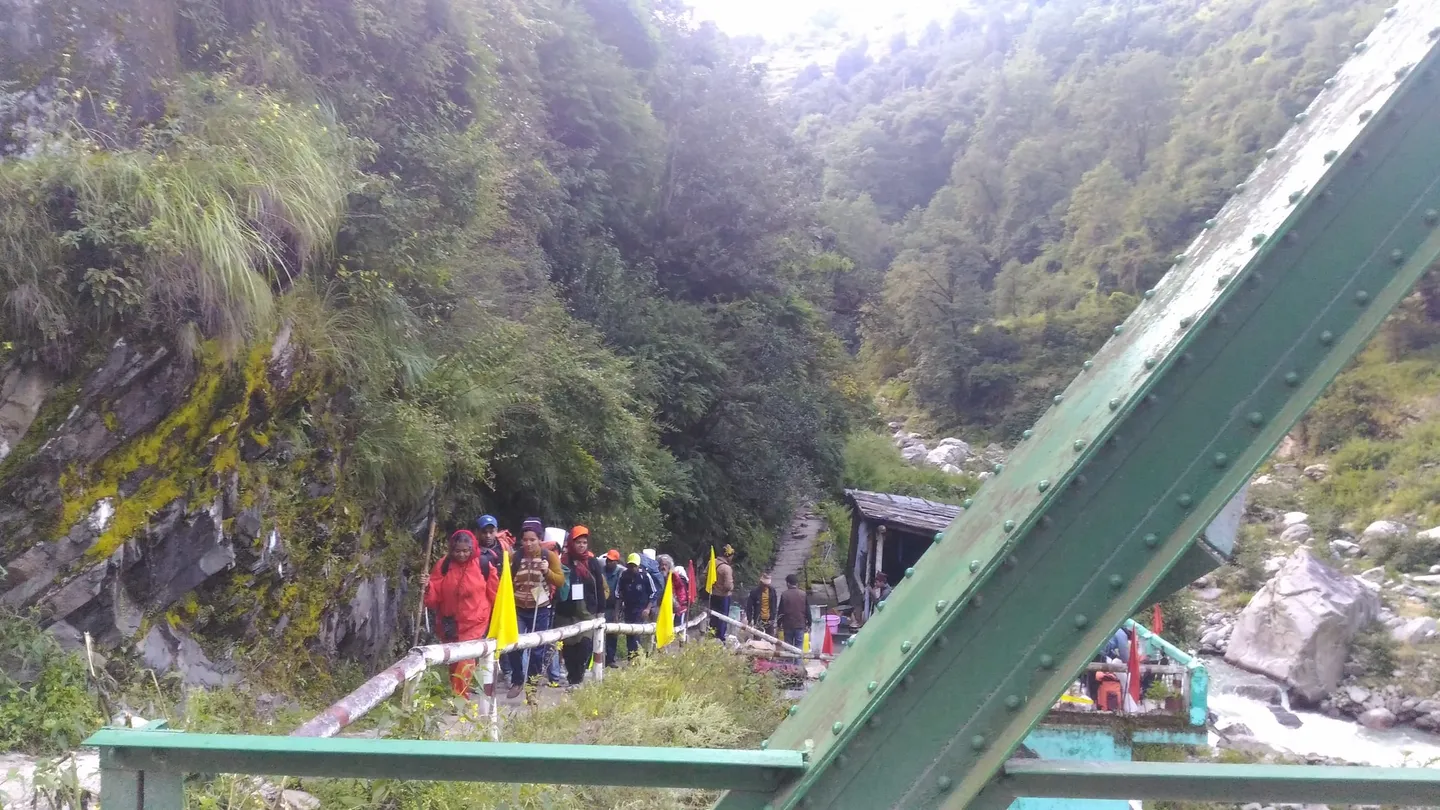 The start of the trail at Hanuman Chatti on a September morning in the monsoon season: a line of walkers with rucksacks, raincoats and yellow flags on a railed path beside a green steel footbridge, with forested slopes behind and a torrent over boulders at the right.