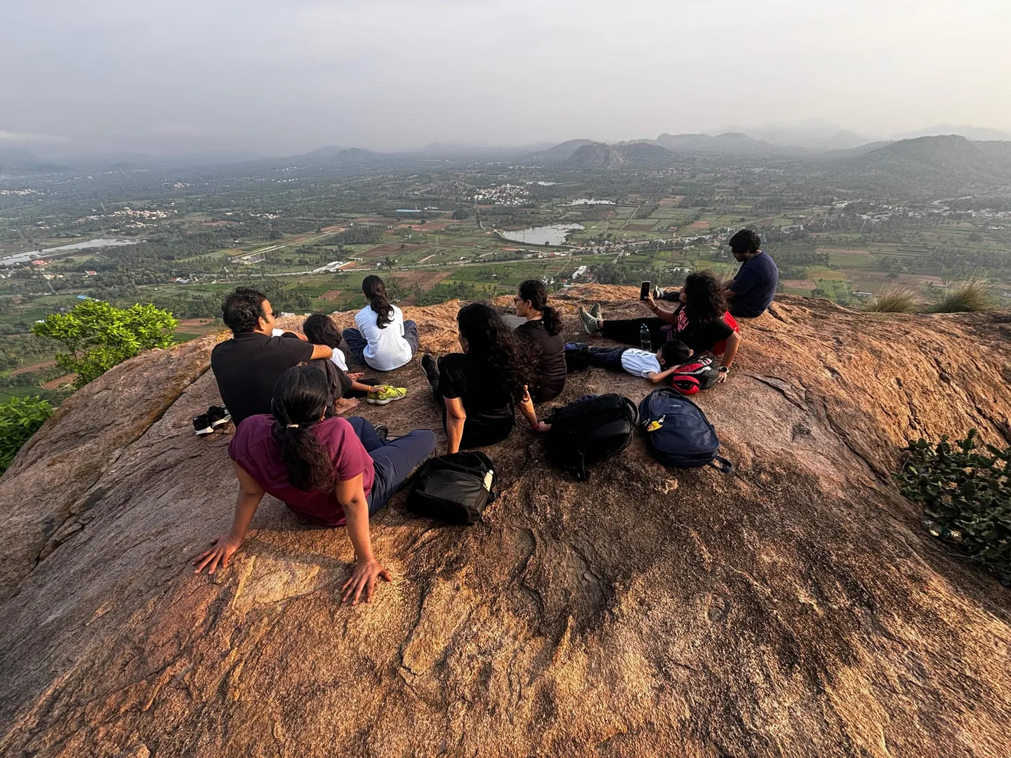 About ten walkers sitting together on the bare granite dome at the top of Achalu Betta at first light, all facing out with their backs to the camera, packs and a water bottle set down on the rock beside them. Below and beyond them the plain opens out for tens of kilometres — paddy squares, coconut groves, two irrigation tanks, a scatter of white and red-roofed villages — with a line of low granite hills along the horizon and soft haze over everything under a pale grey-blue sky. The rock in the foreground is warm orange in the early sun and shows the shallow wandering cracks typical of these domes. Photographed in August, at the end of the monsoon.